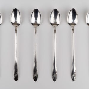 May include: A set of six silver-toned metal spoons arranged in a vertical row. Each spoon features a long, slender handle with a decorative floral engraving at the base and a smooth, oval-shaped bowl at the top, reflecting light on the surface.