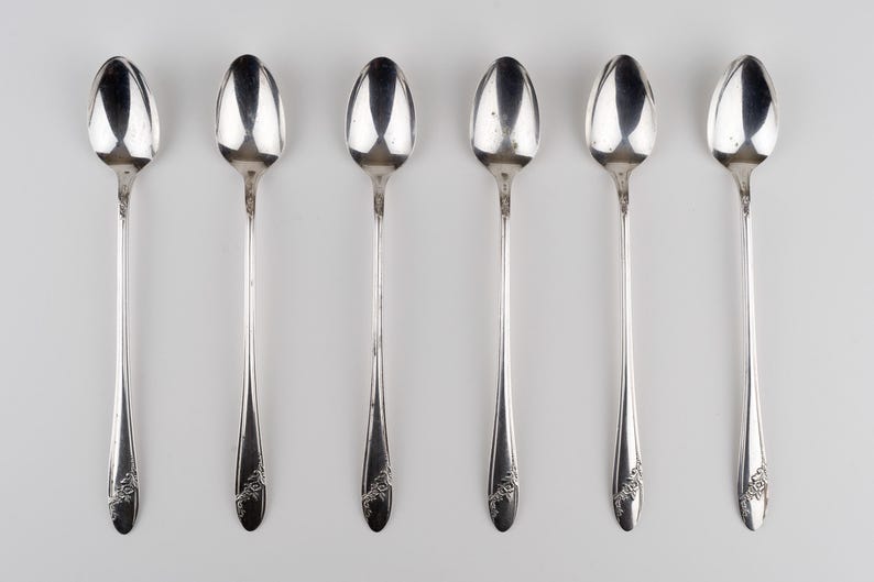 May include: A set of six silver-toned metal spoons arranged in a vertical row. Each spoon features a long, slender handle with a decorative floral engraving at the base and a smooth, oval-shaped bowl at the top, reflecting light on the surface.
