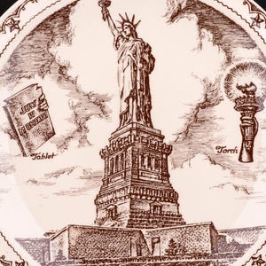 K&ouml;nnte beinhalten: Ein runder wei&szlig;er Keramikteller mit einer detaillierten braunen Tuschezeichnung der Freiheitsstatue. Das Design zeigt eine Tafel mit der Aufschrift JULY IV MDCCLXXVI und eine Fackel, umrahmt von einer dekorativen Bord&uuml;re aus Sternen.