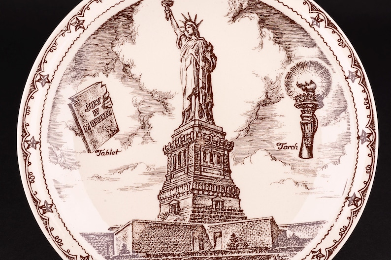K&ouml;nnte beinhalten: Ein runder wei&szlig;er Keramikteller mit einer detaillierten braunen Tuschezeichnung der Freiheitsstatue. Das Design zeigt eine Tafel mit der Aufschrift JULY IV MDCCLXXVI und eine Fackel, umrahmt von einer dekorativen Bord&uuml;re aus Sternen.
