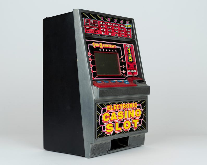 May include: A black and grey electronic casino slot machine game with a digital screen, red control panel, and blue buttons. The front features the text "ELECTRONIC CASINO SLOT" in yellow and red, with a payout table displayed at the top.