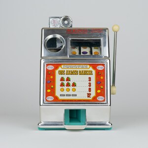 May include: A chrome-finished tabletop slot machine labeled One Armed Banker with a side lever, three spinning reels, and a central display panel featuring fruit symbols, payout values, and the text Medley Mfg Co, Reno, Nevada at the base.
