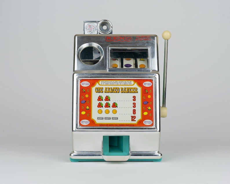 May include: A chrome-finished tabletop slot machine labeled One Armed Banker with a side lever, three spinning reels, and a central display panel featuring fruit symbols, payout values, and the text Medley Mfg Co, Reno, Nevada at the base.