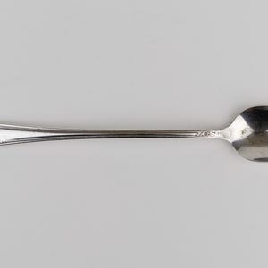 May include: A silver-toned metal spoon featuring an elongated handle with a decorative floral embossed pattern at the tip. The spoon has a smooth, polished finish and a standard oval-shaped bowl, showcasing a classic design with subtle detailing.