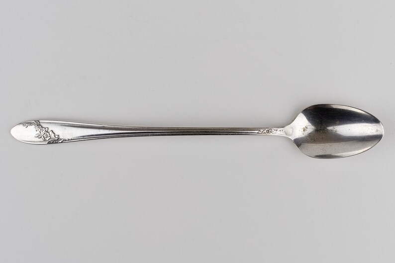 May include: A silver-toned metal spoon featuring an elongated handle with a decorative floral embossed pattern at the tip. The spoon has a smooth, polished finish and a standard oval-shaped bowl, showcasing a classic design with subtle detailing.