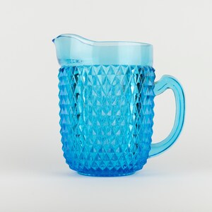May include: A bright blue glass pitcher featuring a prominent diamond-shaped geometric pattern across its exterior surface. The vessel includes a curved handle on the right side and a wide, smooth pouring spout at the top rim for liquid dispensing.