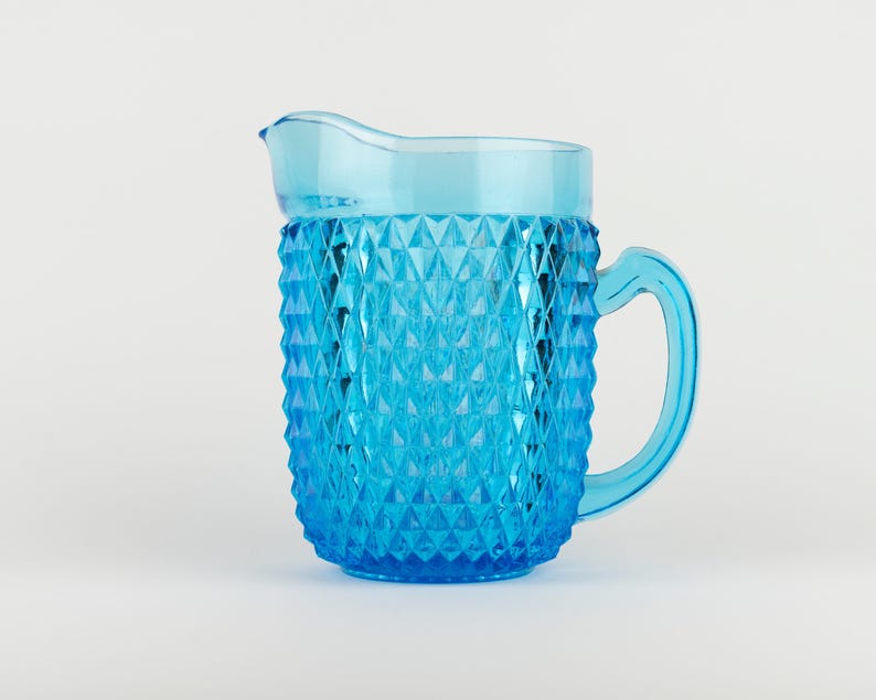 May include: A bright blue glass pitcher featuring a prominent diamond-shaped geometric pattern across its exterior surface. The vessel includes a curved handle on the right side and a wide, smooth pouring spout at the top rim for liquid dispensing.