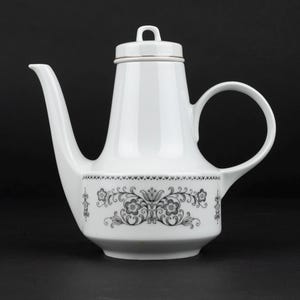 K&ouml;nnte beinhalten: Eine wei&szlig;e Kaffeekanne aus Porzellan mit einem hohen, konischen K&ouml;rper, einem geschwungenen Henkel und einem langen Ausguss. Der untere Teil ist mit einem schwarzen Blumenmuster aus symmetrischen Bl&auml;ttern und Bl&uuml;ten verziert und hat einen Deckel.