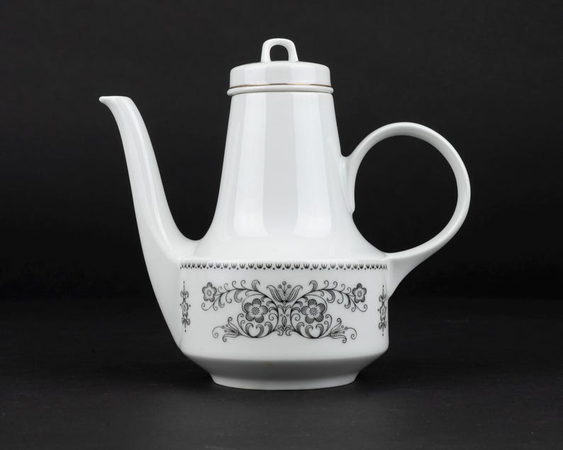 K&ouml;nnte beinhalten: Eine wei&szlig;e Kaffeekanne aus Porzellan mit einem hohen, konischen K&ouml;rper, einem geschwungenen Henkel und einem langen Ausguss. Der untere Teil ist mit einem schwarzen Blumenmuster aus symmetrischen Bl&auml;ttern und Bl&uuml;ten verziert und hat einen Deckel.