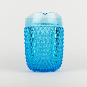 May include: A bright blue translucent pitcher featuring a textured diamond pattern across its exterior surface. The vessel has a smooth, clear rim at the top and a wide, cylindrical body that tapers slightly toward the base, creating a faceted look.