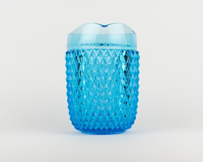 May include: A bright blue translucent pitcher featuring a textured diamond pattern across its exterior surface. The vessel has a smooth, clear rim at the top and a wide, cylindrical body that tapers slightly toward the base, creating a faceted look.