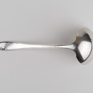 May include: A silver-colored metal ladle with a polished, rounded bowl and a slender handle. The handle features an embossed floral pattern near the tip and includes the stamped text "WM ROGERS MFG CO" and "IS" along the center shaft.