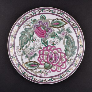 May include: A round ceramic plate featuring a white base with a central floral pattern in shades of magenta, green, and grey. The rim is decorated with a continuous border of small pink flowers and green leaves, accented by thin gold lines.