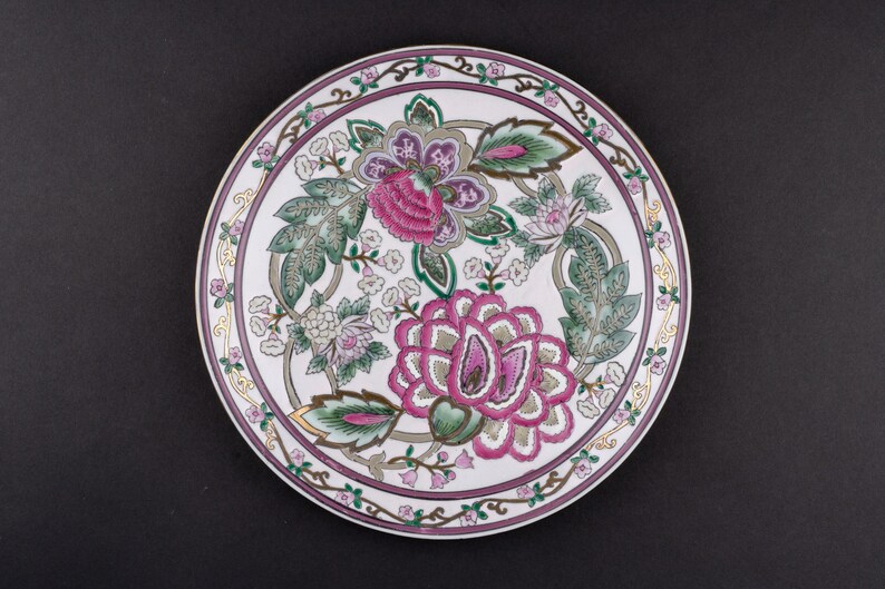 May include: A round ceramic plate featuring a white base with a central floral pattern in shades of magenta, green, and grey. The rim is decorated with a continuous border of small pink flowers and green leaves, accented by thin gold lines.