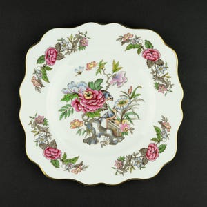 May include: A white square porcelain plate with scalloped edges and a gold rim. The center features a colorful floral arrangement with pink peonies, green leaves, and two small birds. A floral garland border repeats the pink and green motif.