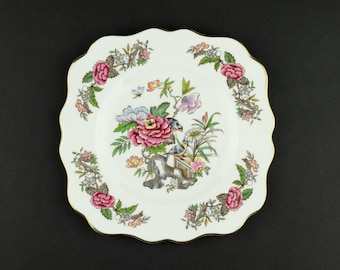 May include: A white square porcelain plate with scalloped edges and a gold rim. The center features a colorful floral arrangement with pink peonies, green leaves, and two small birds. A floral garland border repeats the pink and green motif.
