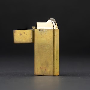 May include: A rectangular brass lighter with a weathered, aged patina finish. The lighter features a hinged lid that is open, revealing a polished gold-tone metal ignition mechanism with a textured flint wheel at the top of the main body.