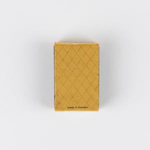 May include: A rectangular matchbox with a mustard yellow cover featuring a dark brown diamond grid pattern. The text "Made in Sweden" is printed in small black letters at the bottom center of the front surface of the matchbox cover.