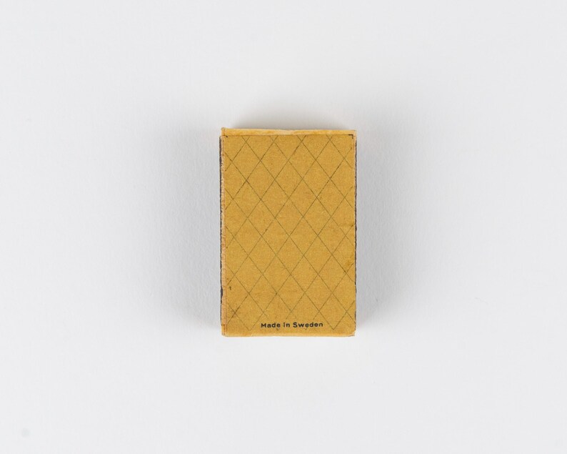 May include: A rectangular matchbox with a mustard yellow cover featuring a dark brown diamond grid pattern. The text "Made in Sweden" is printed in small black letters at the bottom center of the front surface of the matchbox cover.