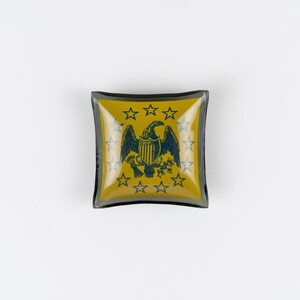 May include: A square glass trinket dish featuring a mustard yellow background with a dark blue eagle emblem at the center. The eagle is surrounded by a circular arrangement of thirteen dark blue stars, all set within a dark-rimmed glass border.