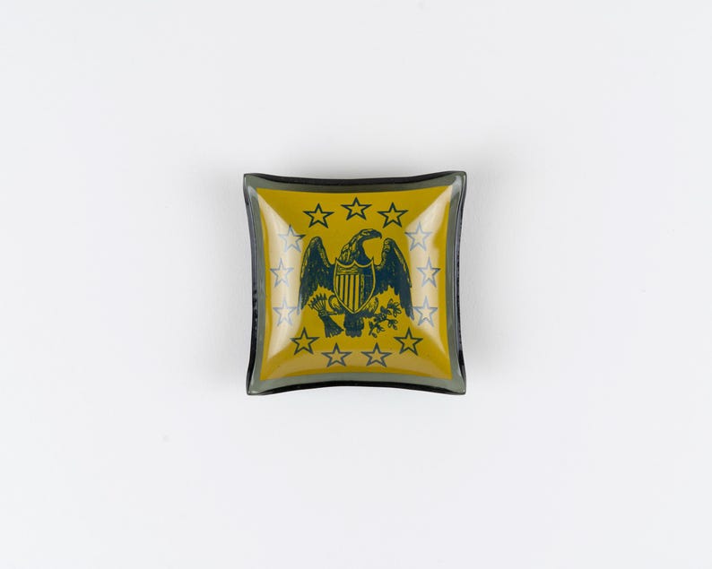 May include: A square glass trinket dish featuring a mustard yellow background with a dark blue eagle emblem at the center. The eagle is surrounded by a circular arrangement of thirteen dark blue stars, all set within a dark-rimmed glass border.