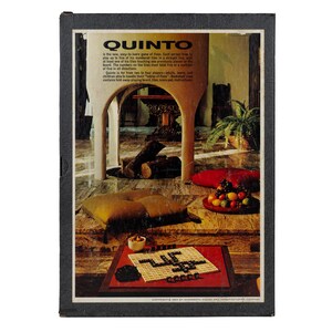 May include: A rectangular board game box titled QUINTO with a black border. The cover features a photograph of a living room with a fireplace, a floor cushion, and a bowl of fruit. In the foreground, a game board with black tiles is displayed.