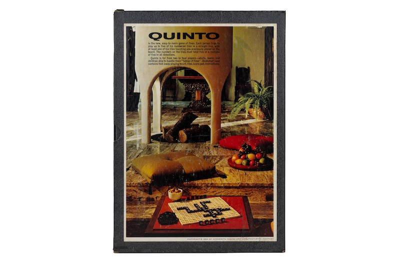 May include: A rectangular board game box titled QUINTO with a black border. The cover features a photograph of a living room with a fireplace, a floor cushion, and a bowl of fruit. In the foreground, a game board with black tiles is displayed.