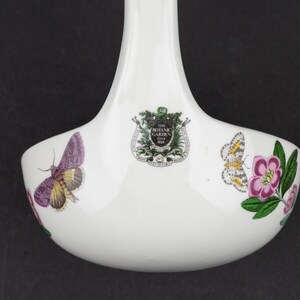 May include: A white ceramic ladle featuring a central crest with the text THE BOTANIC GARDEN 1818. The bowl of the ladle is decorated with colorful illustrations of a purple moth on the left and a patterned butterfly with a pink flower on the right.