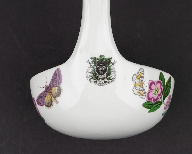May include: A white ceramic ladle featuring a central crest with the text THE BOTANIC GARDEN 1818. The bowl of the ladle is decorated with colorful illustrations of a purple moth on the left and a patterned butterfly with a pink flower on the right.
