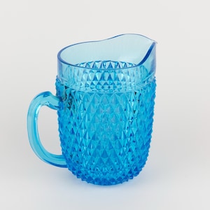 May include: A bright blue glass pitcher featuring a textured diamond pattern across its body. The pitcher has a smooth, clear rim, a curved handle on the side, and a small pouring spout at the top, showcasing a vibrant, translucent finish.