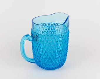 May include: A bright blue glass jug featuring a textured diamond pattern across its body. The jug has a smooth, clear rim, a curved handle on the side, and a small pouring spout at the top, showcasing a vibrant, translucent finish.