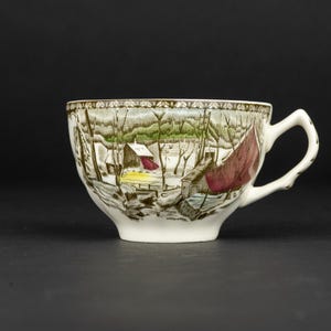 Pu&ograve; includere: Una tazza da t&egrave; in ceramica bianca con una stampa dettagliata di un paesaggio invernale con alberi spogli, un piccolo edificio e un panno rosso drappeggiato su una struttura. Il bordo presenta un motivo decorativo e un manico bianco.