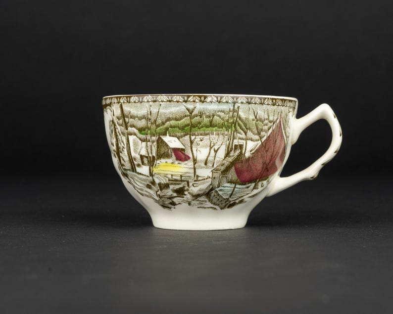 Pu&ograve; includere: Una tazza da t&egrave; in ceramica bianca con una stampa dettagliata di un paesaggio invernale con alberi spogli, un piccolo edificio e un panno rosso drappeggiato su una struttura. Il bordo presenta un motivo decorativo e un manico bianco.