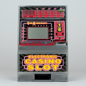 May include: A gray electronic casino slot machine toy featuring a digital screen, red control buttons, and a payout table at the top. The front panel displays the text Electronic Casino Slot in yellow and pink letters with a black background.