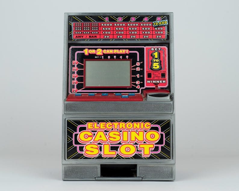 May include: A gray electronic casino slot machine toy featuring a digital screen, red control buttons, and a payout table at the top. The front panel displays the text Electronic Casino Slot in yellow and pink letters with a black background.