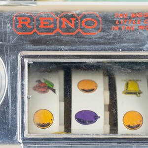 May include: A chrome-finished slot machine panel featuring the red text RENO and THE BIGGEST LITTLE CITY IN THE WORLD. The display window shows three white reels with colorful fruit and bell symbols, including a cherry, orange, plum, and bell.