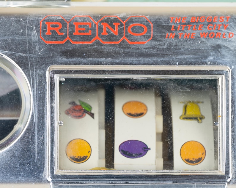 May include: A chrome-finished slot machine panel featuring the red text RENO and THE BIGGEST LITTLE CITY IN THE WORLD. The display window shows three white reels with colorful fruit and bell symbols, including a cherry, orange, plum, and bell.