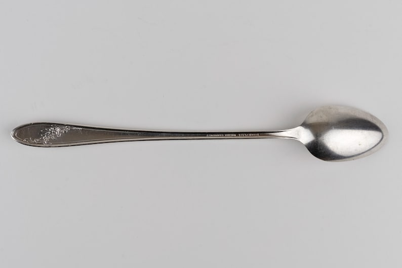 May include: A silver-toned metal spoon featuring a smooth, elongated handle with a decorative floral pattern embossed at the tip. The neck of the spoon displays the text "STAINLESS STEEL" followed by "JAPAN" in small, clear, capital letters.
