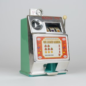 May include: A green and chrome slot machine bank with a side lever and a top coin slot. The front panel features the text "ONE ARMED BANKER" above a payout chart displaying cherries, lemons, and oranges, with a coin tray at the bottom base.