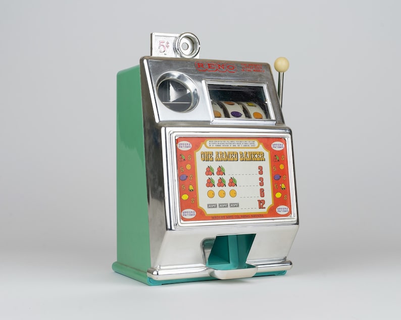 May include: A green and chrome slot machine bank with a side lever and a top coin slot. The front panel features the text "ONE ARMED BANKER" above a payout chart displaying cherries, lemons, and oranges, with a coin tray at the bottom base.