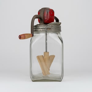 May include: A vintage glass butter churn featuring a square clear glass jar, a metal lid with a red painted gear housing, and a hand crank with a wooden handle. Inside the jar, a wooden paddle mechanism is attached to a central metal rod.