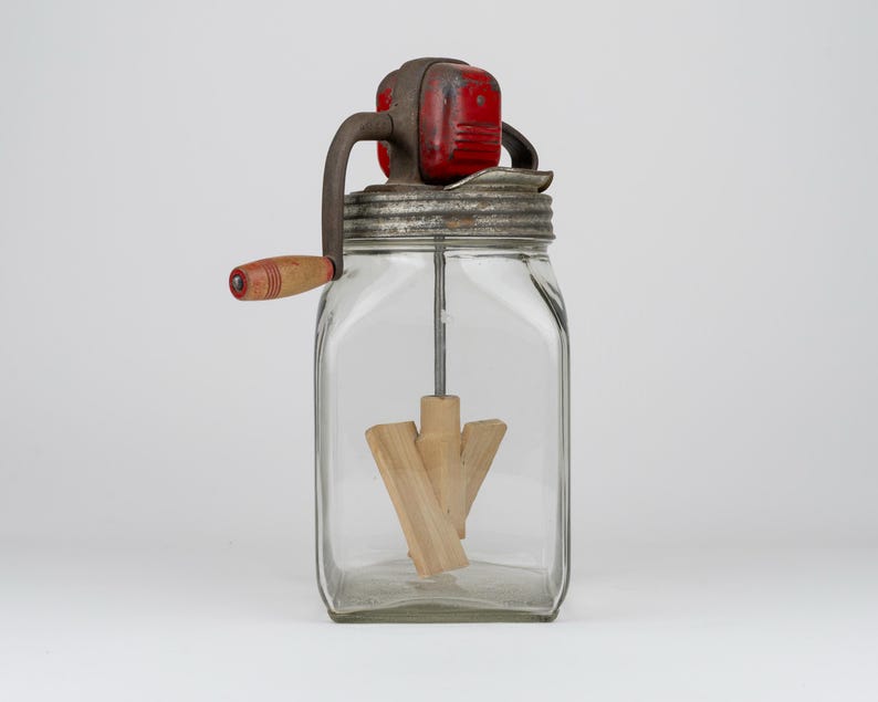 May include: A vintage glass butter churn featuring a square clear glass jar, a metal lid with a red painted gear housing, and a hand crank with a wooden handle. Inside the jar, a wooden paddle mechanism is attached to a central metal rod.