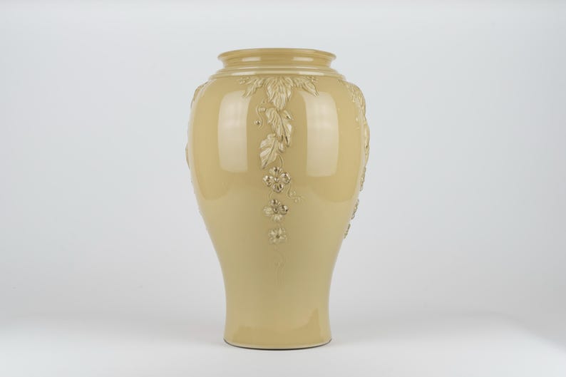 Op de afbeelding: Een glanzende, lichtbeige keramische vaas met een brede, ronde buik en een smalle voet. De buitenkant is voorzien van verhoogde, gegoten reli&euml;fdetails van trapsgewijze bladeren en kleine bloemen met vier bloemblaadjes.