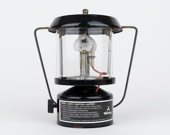 Puede incluir: Una linterna de encendido White Stag modelo H332214 negra con chimenea de cristal transparente, una camisa de tela blanca y un asa de metal. La base presenta una etiqueta negra con texto blanco que detalla las instrucciones de seguridad.