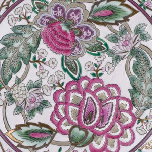 May include: A white ceramic plate featuring a central floral pattern with large pink and purple blossoms, green leaves, and small white flowers. The rim is decorated with a thin purple band and gold filigree accents, framing the detailed botanical design.