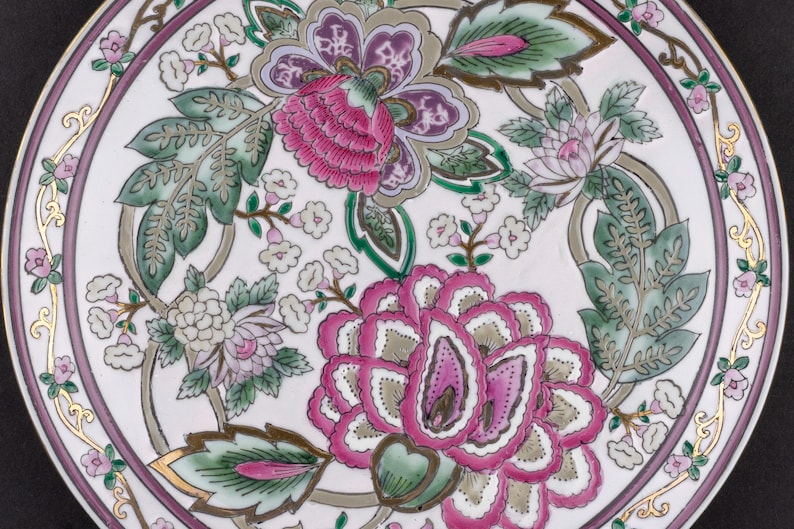 May include: A white ceramic plate featuring a central floral pattern with large pink and purple blossoms, green leaves, and small white flowers. The rim is decorated with a thin purple band and gold filigree accents, framing the detailed botanical design.