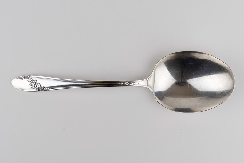 May include: A silver-toned metal spoon featuring a polished oval bowl and a slender handle. The handle is decorated with an embossed floral pattern near the tip and a smaller decorative detail where the handle meets the bowl of the spoon.