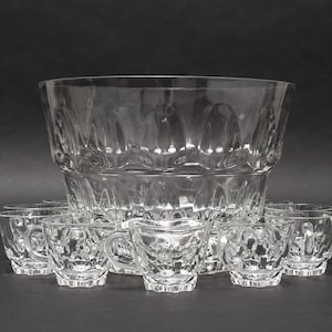 May include: A large clear glass punch bowl with a scalloped rim and a textured pattern, accompanied by a set of matching small glass cups. Each cup features a handle and a repeating embossed design that coordinates with the bowl's exterior.