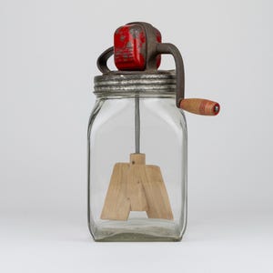 May include: A clear glass butter churn featuring a square-shaped jar, a metal lid, and a red painted gear housing. A hand-crank mechanism with a wooden handle extends from the side, connecting to a wooden paddle agitator inside the jar.