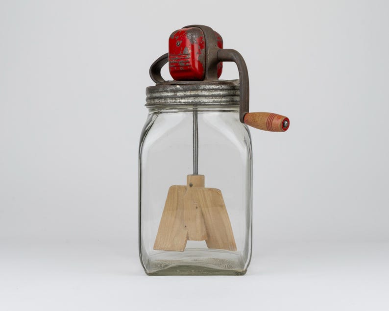 May include: A clear glass butter churn featuring a square-shaped jar, a metal lid, and a red painted gear housing. A hand-crank mechanism with a wooden handle extends from the side, connecting to a wooden paddle agitator inside the jar.